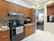 A modern kitchen with wooden cabinets and a stainless steel oven. A kettle sits on the stovetop, and a microwave is on the countertop. The kitchen has a light color scheme with beige tiles and a partial view into a living room with a black and orange color palette.