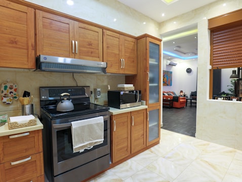 A modern kitchen with wooden cabinets and a stainless steel oven. A kettle sits on the stovetop, and a microwave is on the countertop. The kitchen has a light color scheme with beige tiles and a partial view into a living room with a black and orange color palette.
