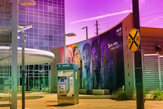 A vibrant urban scene featuring a modern building with reflective glass windows, a ticket machine labeled 'passes' in the foreground, and a large mural on a brick wall displaying diverse faces underlined by the phrase 'You are beautiful.' The sky has an unusual purple hue, creating an artistic atmosphere, and a railroad crossing sign is also visible.