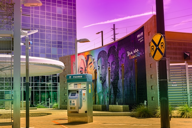 A vibrant urban scene featuring a modern building with reflective glass windows, a ticket machine labeled 'passes' in the foreground, and a large mural on a brick wall displaying diverse faces underlined by the phrase 'You are beautiful.' The sky has an unusual purple hue, creating an artistic atmosphere, and a railroad crossing sign is also visible.