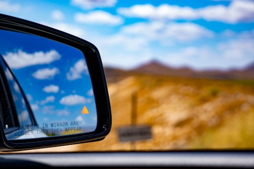 A close-up view of a car's side mirror reflecting a clear sky with scattered clouds. An orange warning triangle is visible on the mirror, along with the text 'OBJECTS IN MIRROR ARE CLOSER THAN THEY APPEAR'. The background features a blurred landscape of hills under a bright blue sky.