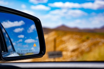 A close-up view of a car's side mirror reflecting a clear sky with scattered clouds. An orange warning triangle is visible on the mirror, along with the text 'OBJECTS IN MIRROR ARE CLOSER THAN THEY APPEAR'. The background features a blurred landscape of hills under a bright blue sky.