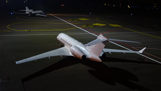 Two private jets are parked on a dimly lit runway at night, with one jet in the foreground and another in the background. The scene is illuminated by artificial light, reflecting off the sleek surfaces of the aircraft. The ground is marked with various lines and labels typical of an airport taxiway.