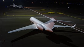 Two private jets are parked on a dimly lit runway at night, with one jet in the foreground and another in the background. The scene is illuminated by artificial light, reflecting off the sleek surfaces of the aircraft. The ground is marked with various lines and labels typical of an airport taxiway.