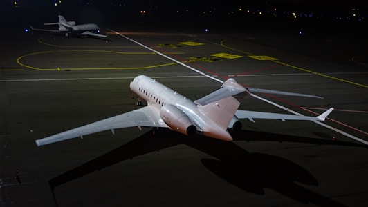 Two private jets are parked on a dimly lit runway at night, with one jet in the foreground and another in the background. The scene is illuminated by artificial light, reflecting off the sleek surfaces of the aircraft. The ground is marked with various lines and labels typical of an airport taxiway.