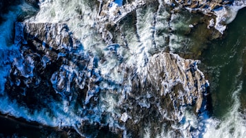Aerial view of a dynamic river scene featuring powerful white rapids coursing over jagged rocks. The whitewater contrasts sharply with the dark, smooth surfaces of the rock formations and the swirling green and blue hues of the water, creating a sense of motion and energy.