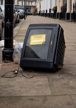 A large, old-style CRT television is discarded on a sidewalk next to a streetlight pole. The television has a sign on it indicating that illegal fly-tipping has occurred. Nearby, there are plastic bags filled with trash. The scene is set in an urban area with parked cars lining the street and residential buildings in the background.