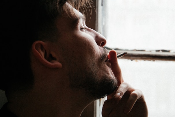 A person with a contemplative expression holds a lit cigarette close to their lips. Soft natural light comes through the window, casting gentle shadows across their face.