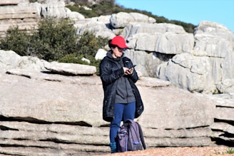 A person stands outdoors on rocky terrain, wearing a red cap, a dark jacket, and blue pants. They are focused on a smartphone in their hands. A backpack is placed on the ground next to them. The background features large rock formations and some greenery.