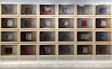 A series of framed displays mounted on a wall, each containing a folded, neatly arranged piece of textile or clothing. The backgrounds of the frames are uniformly textured, creating a cohesive aesthetic. The textiles vary in color and pattern, showing an assortment of styles and materials.