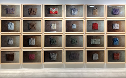 A series of framed displays mounted on a wall, each containing a folded, neatly arranged piece of textile or clothing. The backgrounds of the frames are uniformly textured, creating a cohesive aesthetic. The textiles vary in color and pattern, showing an assortment of styles and materials.