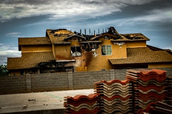 A two-story house with significant fire damage on the roof and upper facade, with a partially collapsed structure. Charred and blackened areas are visible where the fire has burned through. In the foreground, stacks of roofing tiles are piled up, while a concrete wall separates the building from the area where the tiles are stored. The sky is cloudy, casting a somber atmosphere over the scene.