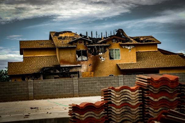 A two-story house with significant fire damage on the roof and upper facade, with a partially collapsed structure. Charred and blackened areas are visible where the fire has burned through. In the foreground, stacks of roofing tiles are piled up, while a concrete wall separates the building from the area where the tiles are stored. The sky is cloudy, casting a somber atmosphere over the scene.