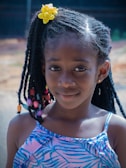 A young girl with braided hair adorned with colorful beads and a bright yellow flower accessory. She is wearing a blue patterned dress and smiles gently at the camera, with a blurred outdoor background.