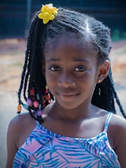 A young girl with braided hair adorned with colorful beads and a bright yellow flower accessory. She is wearing a blue patterned dress and smiles gently at the camera, with a blurred outdoor background.
