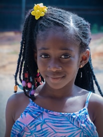A young girl with braided hair adorned with colorful beads and a bright yellow flower accessory. She is wearing a blue patterned dress and smiles gently at the camera, with a blurred outdoor background.