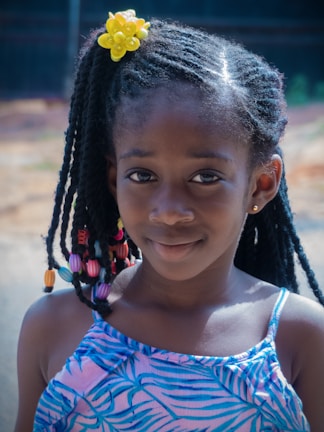 A young girl with braided hair adorned with colorful beads and a bright yellow flower accessory. She is wearing a blue patterned dress and smiles gently at the camera, with a blurred outdoor background.