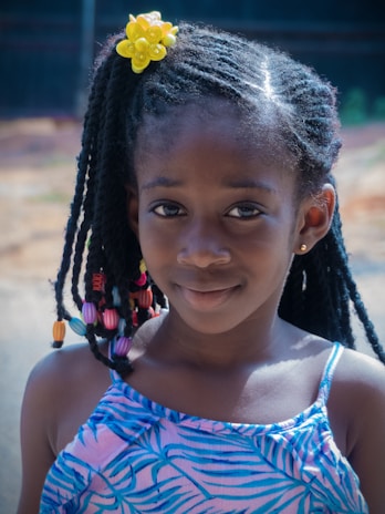A young girl with braided hair adorned with colorful beads and a bright yellow flower accessory. She is wearing a blue patterned dress and smiles gently at the camera, with a blurred outdoor background.