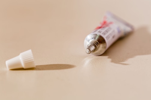 A small, silver metallic tube with a twist-off cap lies on a light-colored surface. The cap is detached and placed next to the tube, which is in focus towards the foreground.