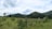 A serene countryside landscape features green rolling hills covered with dense trees under a partly cloudy sky. In the foreground, there is a wooden fence and a grass field with scattered bushes and small trees. A couple of small houses with green roofs are visible in the distance.