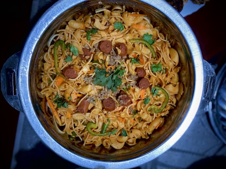 A metallic pot is filled with a dish consisting of spiral pasta mixed with sliced sausages, green peppers, and garnished with fresh cilantro. The pasta is lightly coated with a red sauce, possibly tomato-based, and there are visible slices of cooked meat and vegetables integrated throughout the dish.