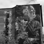 A large mural is painted on the side of a brick building, depicting an elderly man with a beard holding flowers. The artwork is in grayscale, creating a striking effect against the building's red brick. On the left side, several smaller faces and a figure climbing a ladder are also painted, adding complexity to the mural.