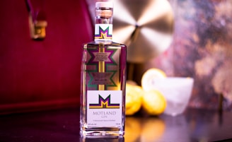 A bottle of Motland Gin is prominently displayed on a reflective surface. The label features a colorful geometric design with a bold M at the center. In the background, there are two lemons and a glass with a lemon slice on the rim. The setting includes a blurred, elegant backdrop with warm, reddish hues.