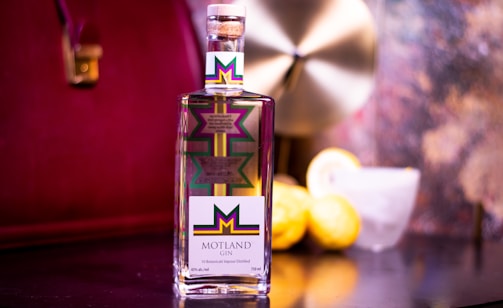 A bottle of Motland Gin is prominently displayed on a reflective surface. The label features a colorful geometric design with a bold M at the center. In the background, there are two lemons and a glass with a lemon slice on the rim. The setting includes a blurred, elegant backdrop with warm, reddish hues.