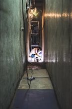 An individual is crouched down in a narrow, dimly lit hallway, possibly performing some form of welding or electrical work. The narrow space is lined with grey walls and scattered cables. The person is illuminated by a bright light source, emphasizing the activity. Loose wires and a small blue and white object are seen in the foreground, leading towards the person working.