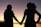 Two people hold hands against a scenic sunset backdrop. Their silhouettes are clearly defined as they face each other, suggesting a moment of connection and intimacy.
