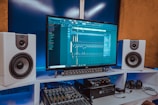 A modern music production setup featuring a large monitor displaying digital audio software. Two white speakers are positioned on either side. Below the monitor is audio equipment, including a sound mixer, and a pair of headphones rests on a desk.