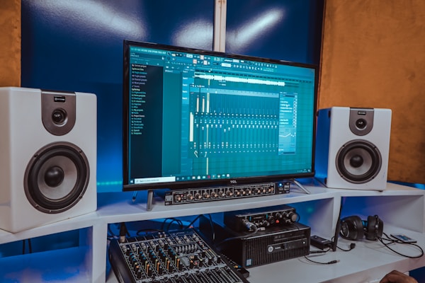 A modern music production setup featuring a large monitor displaying digital audio software. Two white speakers are positioned on either side. Below the monitor is audio equipment, including a sound mixer, and a pair of headphones rests on a desk.