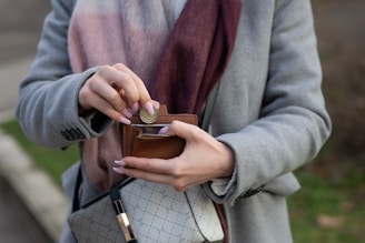 A person dressed in a gray coat and a burgundy scarf is holding a small brown wallet. They are inserting a coin into the wallet. The person has manicured nails and is carrying a light-colored designer handbag with a patterned design and a gold accent.