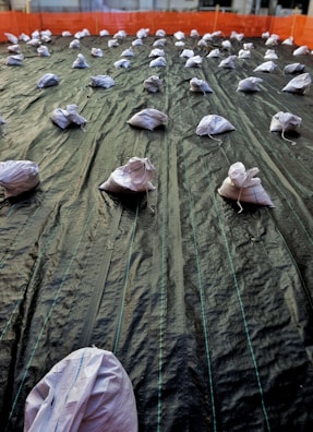 A large area is covered with a dark tarp material, held down by multiple white sandbags. The tarp has visible seams and lines running parallel across its length. The sandbags are evenly distributed in rows and the background features an orange safety barrier surrounding the area.