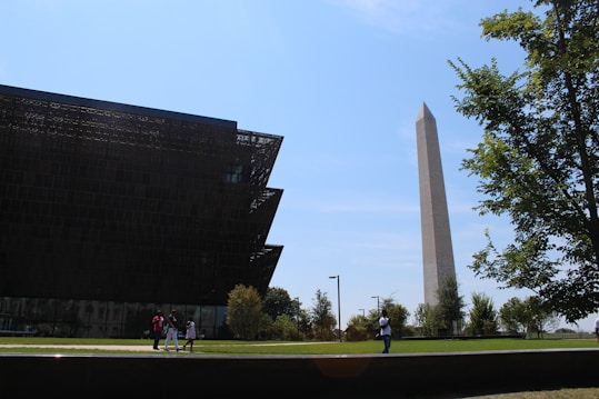 A modern museum building with intricate lattice-like architecture stands beside a tall, iconic obelisk monument. The sky is clear and blue. There are a few people walking and taking photos on the green lawn that surrounds the structures, and trees are visible, adding some greenery to the scene.