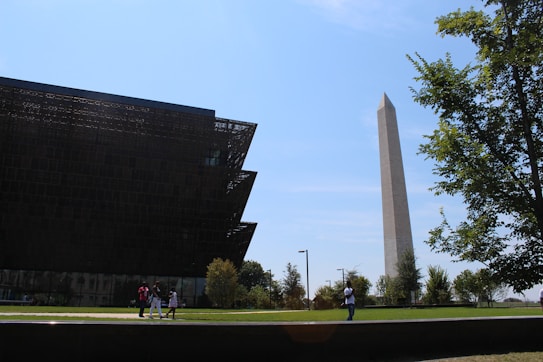 A modern museum building with intricate lattice-like architecture stands beside a tall, iconic obelisk monument. The sky is clear and blue. There are a few people walking and taking photos on the green lawn that surrounds the structures, and trees are visible, adding some greenery to the scene.