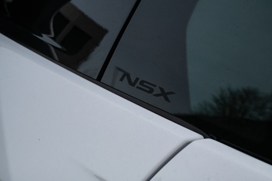 A close-up view of a car with a white body and a dark-colored window. The logo 'NSX' is visible on the window, indicating a vehicle model. The surrounding area appears dimly lit, possibly at dusk or in a shaded area.