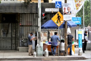 A street food stall is set up on a sidewalk with a blue canopy, surrounded by city infrastructure and signs. Three people are present: one reads a newspaper, while two are preparing or serving food. The stall includes large containers likely used for beverages or food storage. An assortment of signs and posters, some political, are attached to the pole and surrounding area.