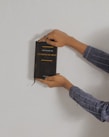 A person holding a black book against a plain background. The book is titled 'The Rules of Entrepreneurship' and is grasped with both hands. The person is wearing a plaid shirt with long sleeves and has a ring on one finger.
