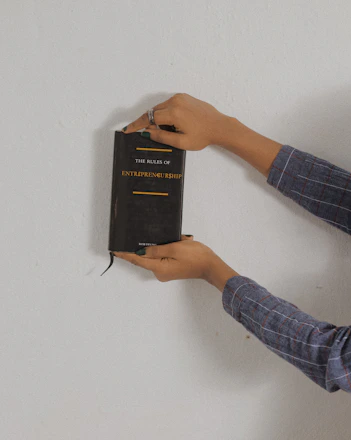 A person holding a black book against a plain background. The book is titled 'The Rules of Entrepreneurship' and is grasped with both hands. The person is wearing a plaid shirt with long sleeves and has a ring on one finger.
