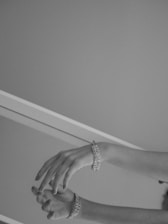 A pair of elegantly posed hands adorned with intricate bracelets are set against a minimalist background. The image is captured in grayscale, highlighting the delicate structure and positioning of the fingers. The lighting creates subtle shadows, enhancing the texture of the bracelets.