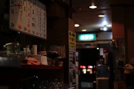 A dimly lit interior of what appears to be a cozy Japanese restaurant. The counter is cluttered with dishes, glasses, bottles, and a menu written in Japanese. There is an illuminated exit sign in the background along with soft ambient lighting from overhead lamps. The atmosphere feels intimate and slightly nostalgic, characteristic of small, traditional eateries.
