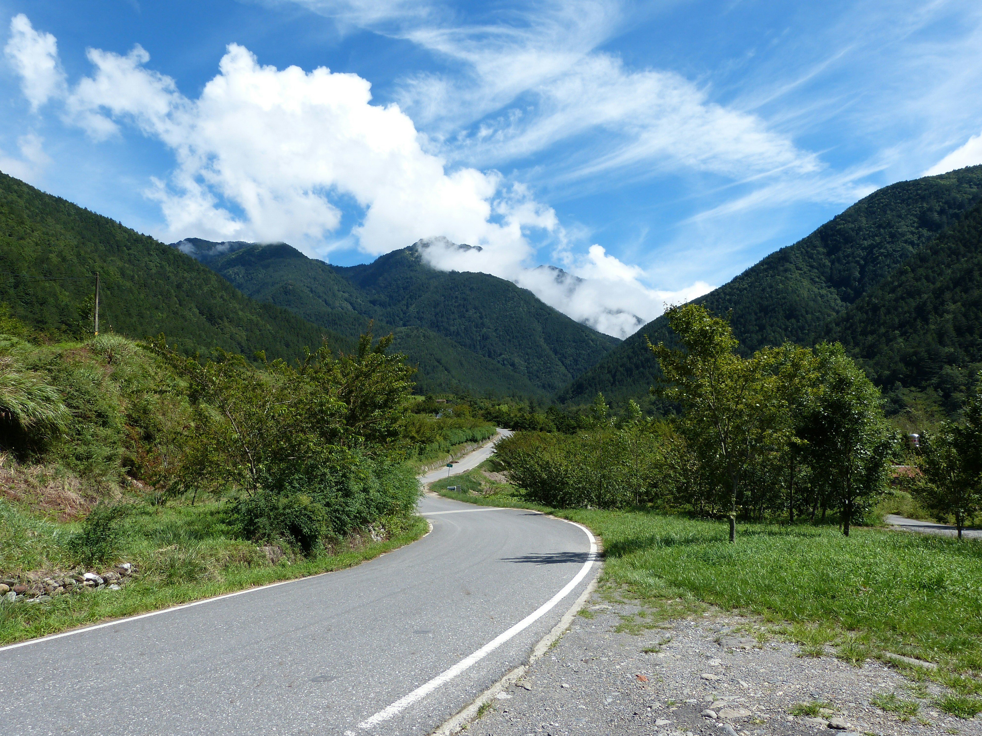 gray concrete road between green grass field and green mountains under blue and white cloudy sky