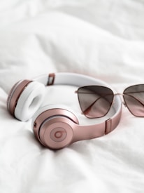 A pair of rose gold and white over-ear headphones is placed on a soft, white fabric. Beside the headphones, there is a pair of stylish sunglasses with tinted lenses. The arrangement suggests a casual and relaxed atmosphere.