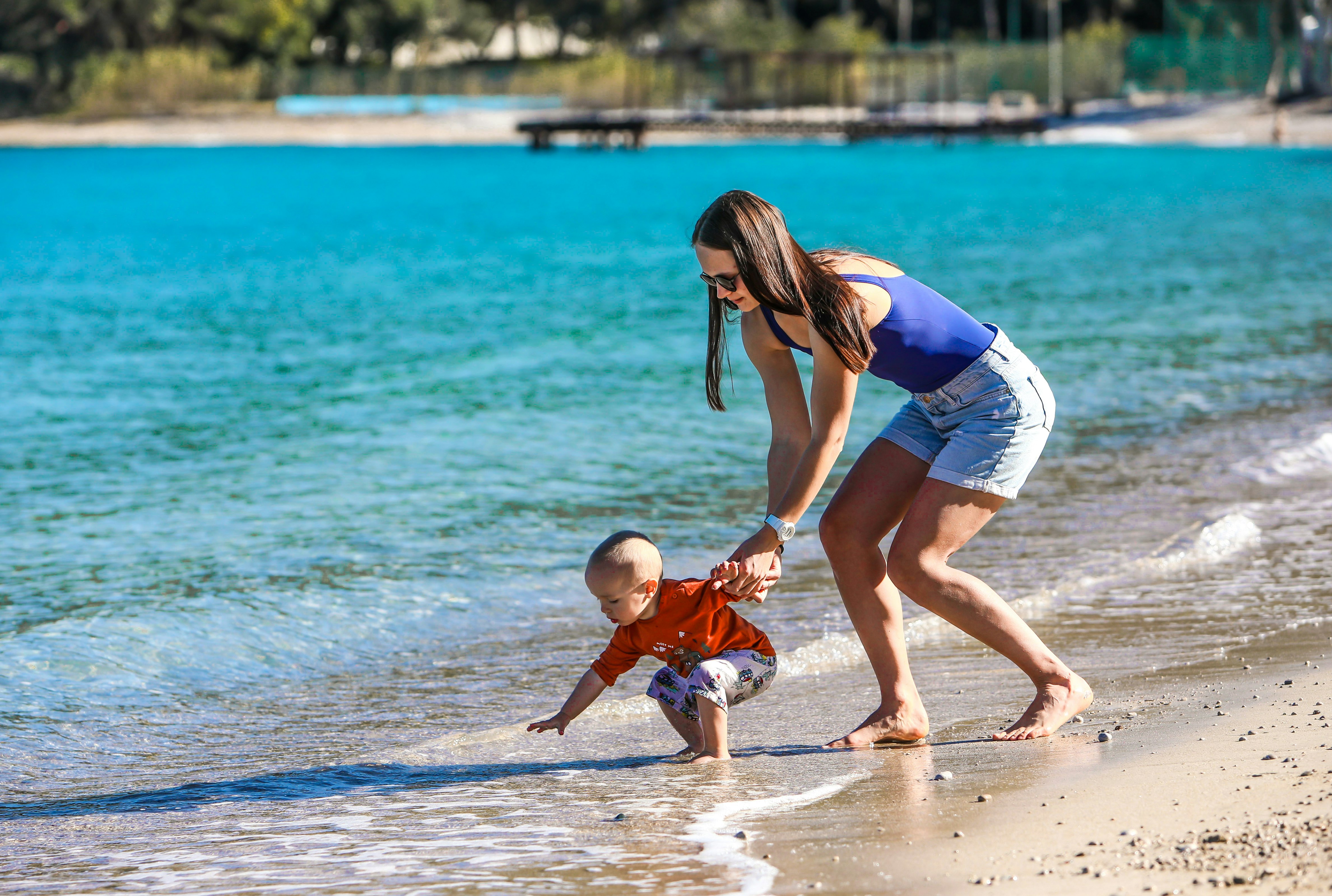 woman in blue and white tank top holding baby in white onesie on beach during daytime