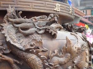 Intricately carved dragon sculpture integrated into a large, ornate vessel with detailed scales and dynamic posture. The surface is adorned with traditional motifs and Chinese characters, suggesting cultural or religious significance. Background hints at an urban setting with blurred colorful signs and buildings, creating a contrast between the ancient art and modern environment.