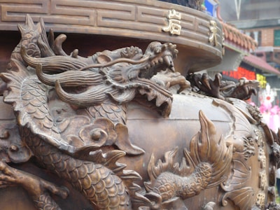 Intricately carved dragon sculpture integrated into a large, ornate vessel with detailed scales and dynamic posture. The surface is adorned with traditional motifs and Chinese characters, suggesting cultural or religious significance. Background hints at an urban setting with blurred colorful signs and buildings, creating a contrast between the ancient art and modern environment.