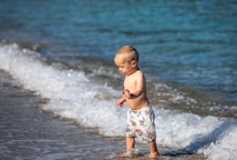 A toddler is standing in shallow water at the edge of the sea, dressed in colorful shorts with a pattern. The child appears to be playing with the water as small waves come in. The background features clear, blue water.