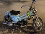 A vintage motorbike with light blue body paint and a single seat. It features a rugged off-road tire at the front and a metal luggage rack at the back. A plastic water bottle is attached to the frame near the handlebars. The surroundings include a road surface with scattered leaves.