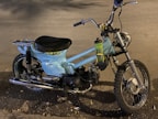 A vintage motorbike with light blue body paint and a single seat. It features a rugged off-road tire at the front and a metal luggage rack at the back. A plastic water bottle is attached to the frame near the handlebars. The surroundings include a road surface with scattered leaves.